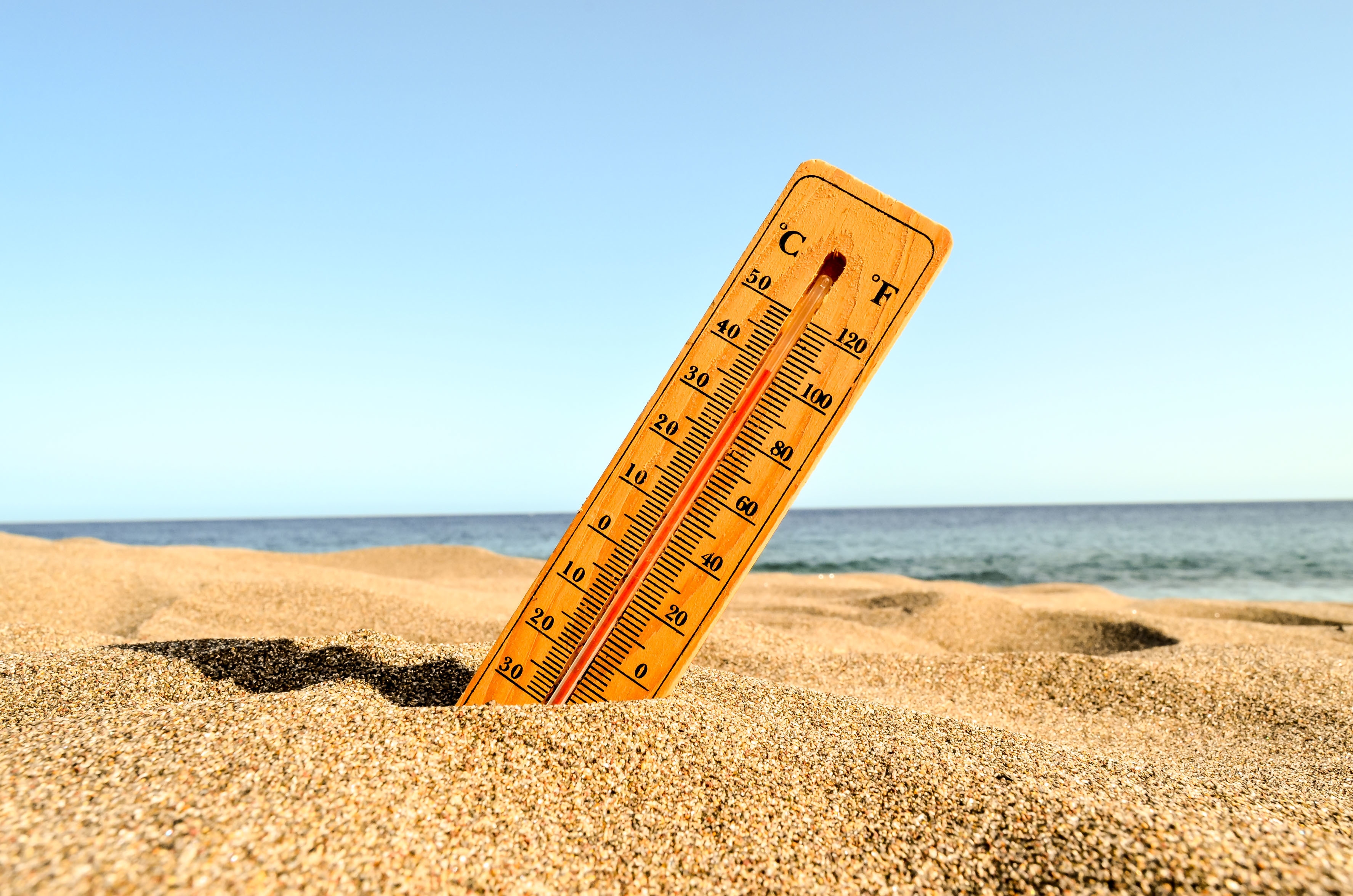 Thermomètre en bois planté dans le sable d'une plage ensoleillée, affichant une température élevée, avec la mer en arrière-plan sous un ciel bleu dégagé.